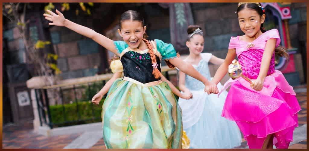 Three smiling young girls in colorful princess dresses hold hands and play together outdoors at a birthday celebration