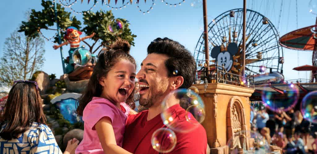 A father joyfully lifts his daughter at an amusement park with rides and bubbles in the background on a bright sunny day