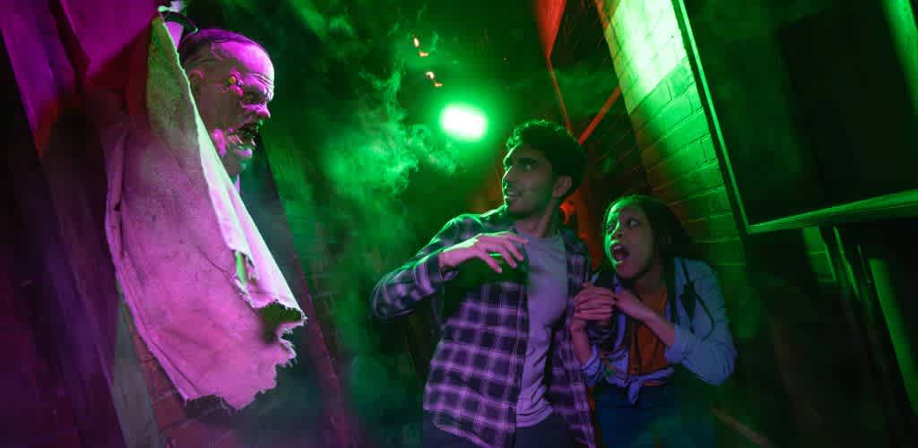 Two people are frightened by a ghostly figure with a sinister expression in a haunted house with green and purple lighting.