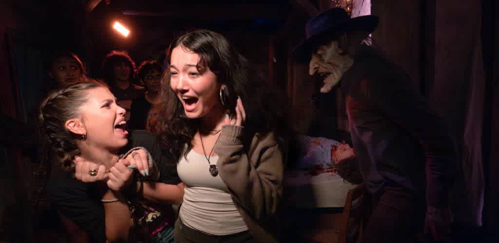 Two young women look frightened and distressed as a scary clown in a dark room with red lighting looms nearby.