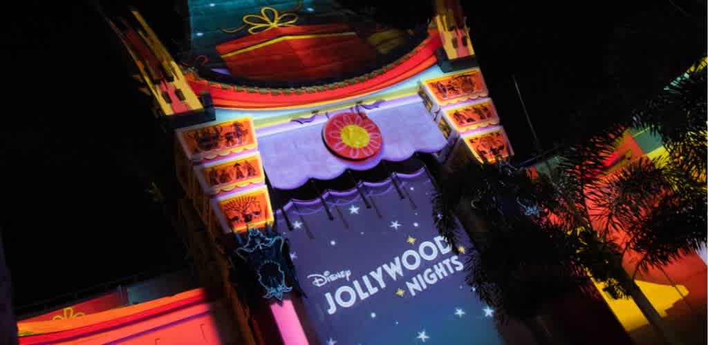 Brightly lit Disney Jollywood Nights marquee sign with colorful decorations and palm trees during nighttime.