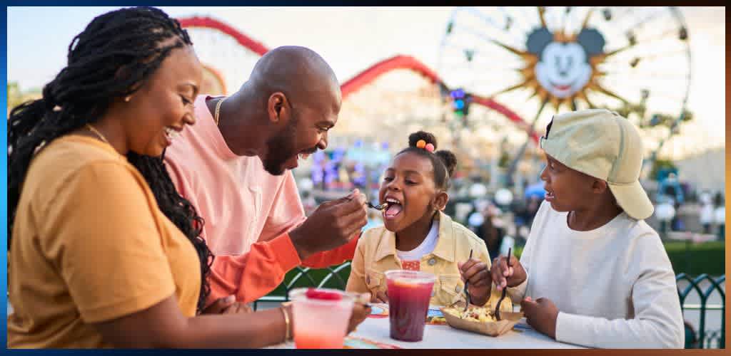 A family enjoys ice cream together outdoors at an amusement park with a Ferris wheel in the background and a Mickey Mouse face decoration.