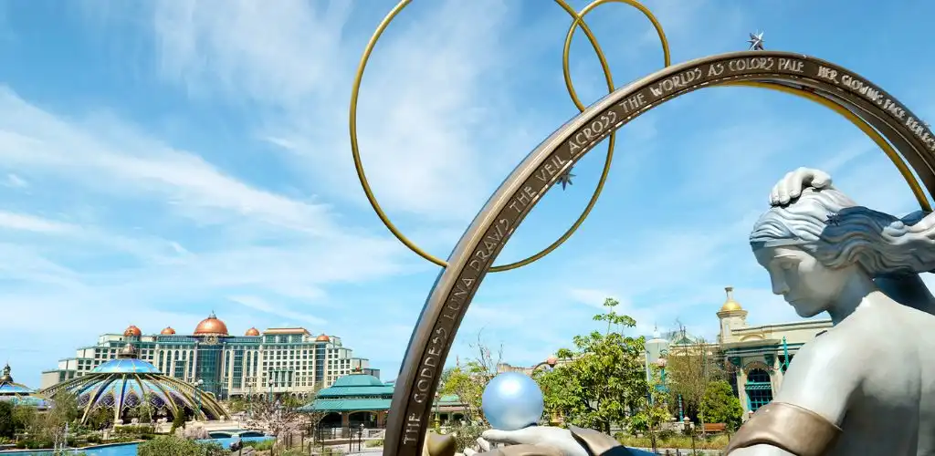 Statue of a woman holding a sphere with large golden rings and a colorful theme park in the background under a blue sky