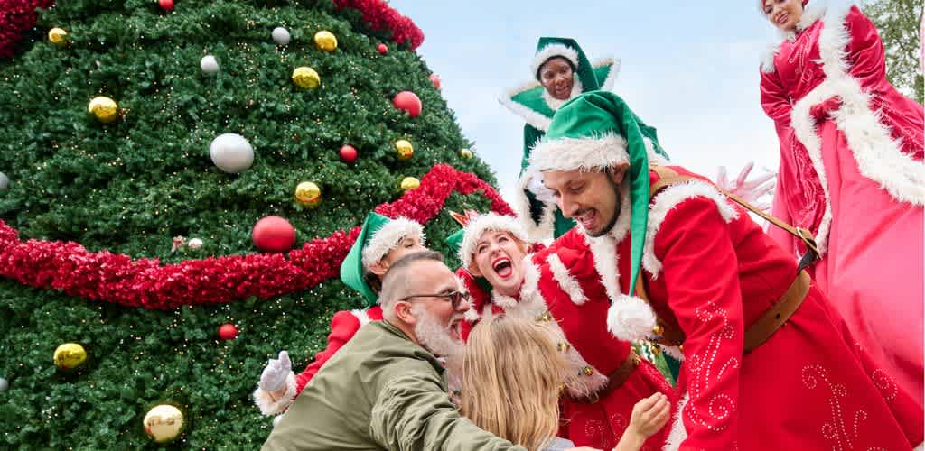 A group of people in festive costumes celebrating outdoors next to a decorated Christmas tree with ornaments and garland.