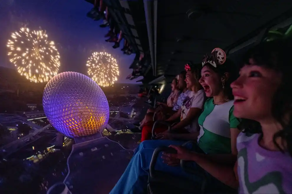 Guests take flight on Soarin’ at EPCOT, soaring high above a glowing Spaceship Earth as fireworks light up the night sky, creating a breathtaking and immersive experience. 