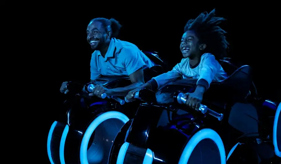 Father and child race through the digital frontier on a high-speed lightcycle, glowing with blue neon lights on TRON at Magic Kingdom.