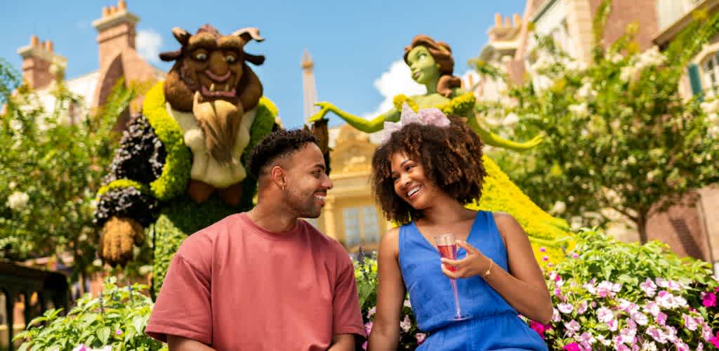 Two people smile and talk outdoors with large colorful fairy tale characters in the background, surrounded by blooming flowers on a bright sunny day.