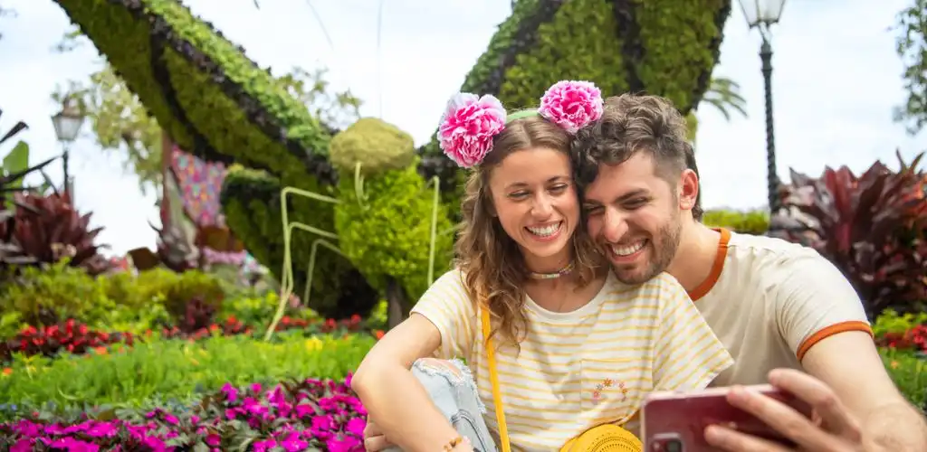 A happy couple takes a selfie outdoors among colorful flowers and greenery, smiling joyfully with a large moss-covered art installation in the background.