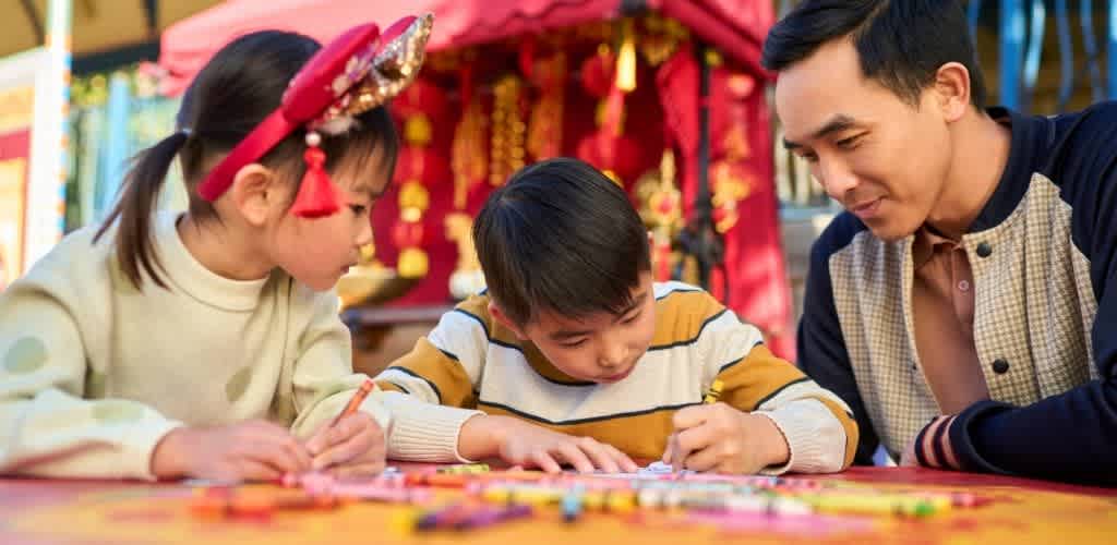 A man and two children engaging in a colorful craft activity outdoors with decorated tents in the background.