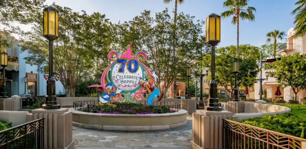 A colorful display in a courtyard features '70 Celebrate! Be Happy' surrounded by trees, street lamps, and decorative fencing.