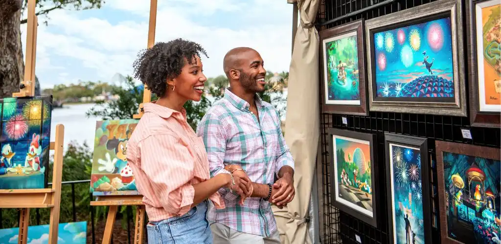 A smiling couple viewing colorful outdoor artwork at an art festival by a river with trees in the background.