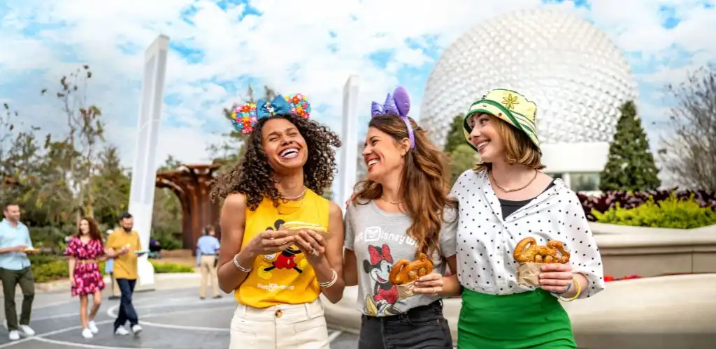 Three women smiling and holding pretzels outdoors at a Disney location with the Epcot geodesic sphere in the background