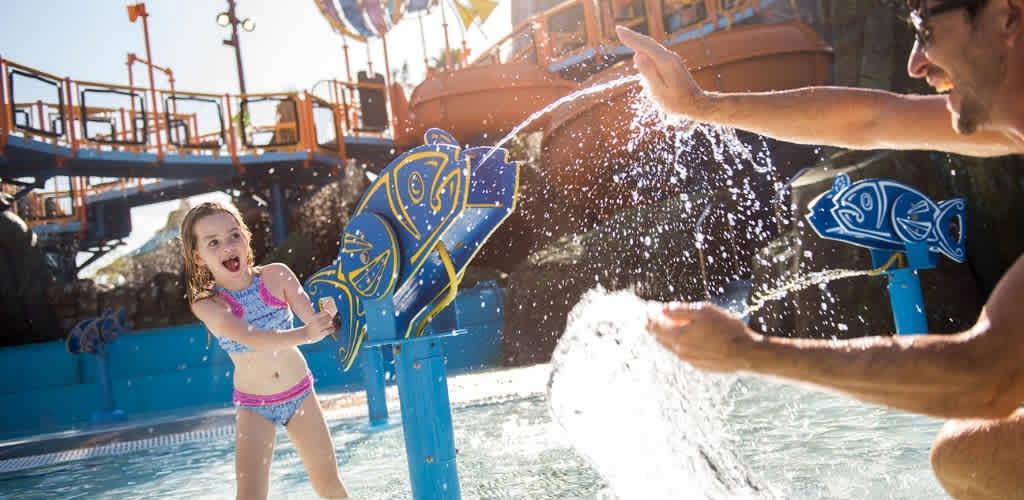 A young girl enjoying water play at a themed water park with an adult splashing water in the background.