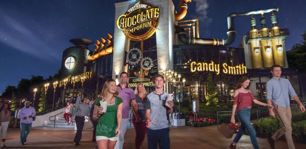 Visitors walking outside a themed chocolate and candy store at night with illuminated signs and industrial design elements.