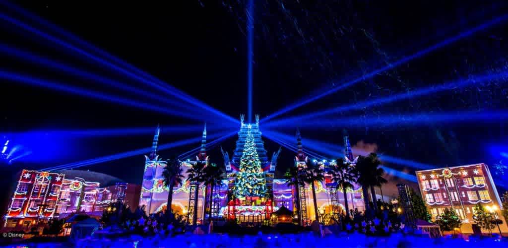 Nighttime holiday scene with colorful lights, a decorated Christmas tree, palm trees, and light projections on buildings and the sky.