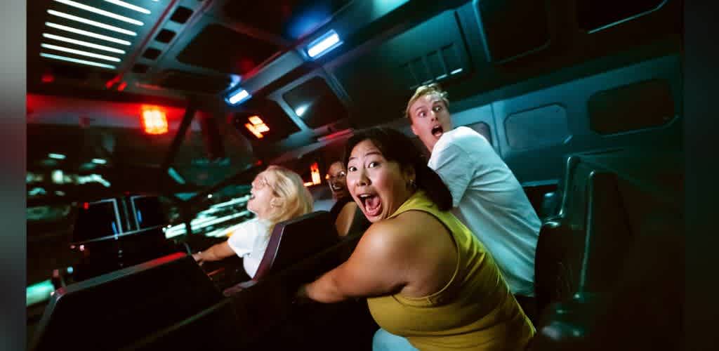 A group of people in a vehicle with excited facial expressions, illuminated by colorful lights, experiencing a thrilling ride or simulation inside a futuristic setting.