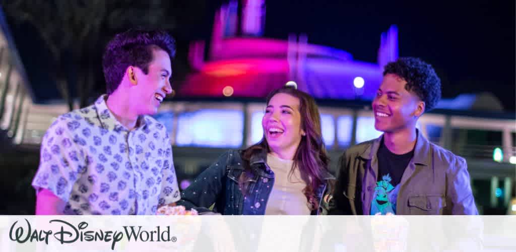 A group of three friends laughing and enjoying snacks together under vibrant nighttime lights at Magic Kingdom Park during Disney After Hours