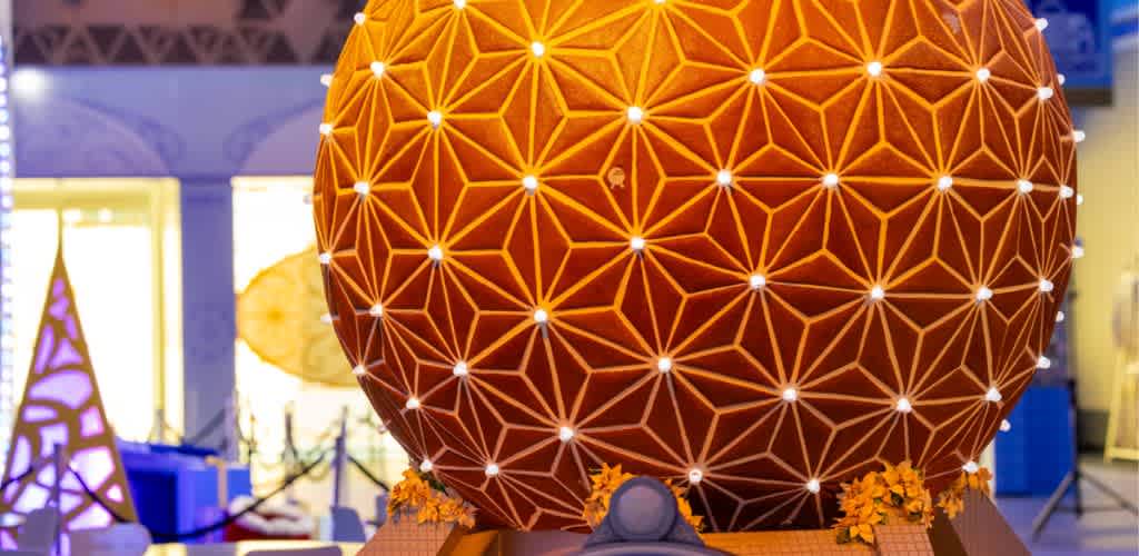 Large spherical geometric lantern with a warm orange glow and intricate white pattern, decorated with yellow flowers at its base in an indoor setting.