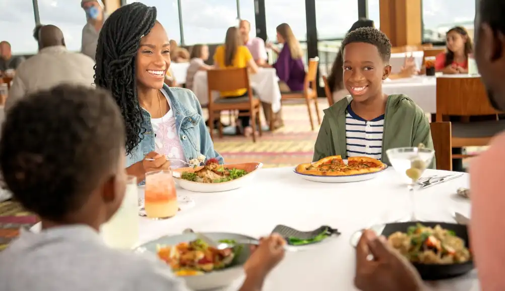 A family enjoys a delicious meal together in an elegant Disney restaurant, sharing smiles and laughter while dining with a scenic view.