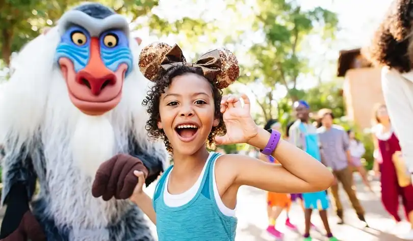 A joyful young girl wearing sparkly Minnie ears shares a fun moment with Rafiki at Disney’s Animal Kingdom, surrounded by vibrant nature and lively atmosphere.