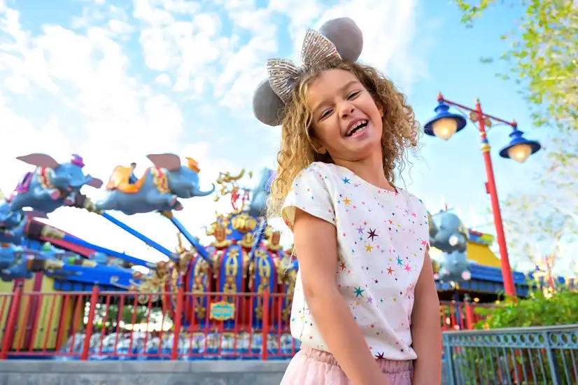 A smiling young girl wearing sparkly Mickey ears enjoys a magical day at Magic Kingdom Park, with Dumbo the Flying Elephant soaring behind her in Fantasyland.