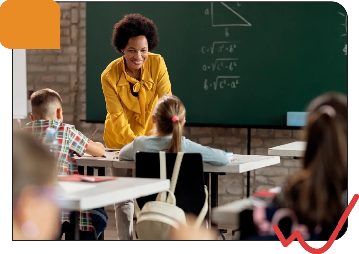 A smiling female teacher in a yellow jacket engaging young students in a classroom with a chalkboard background and educational materials.