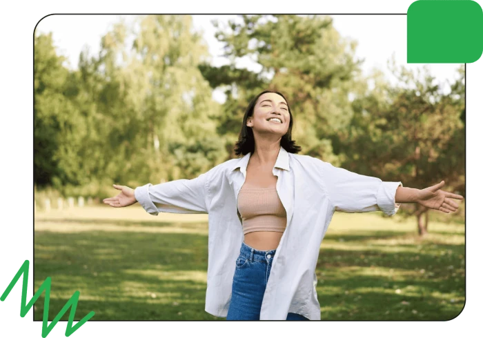 A woman standing outdoors with arms outstretched enjoying a sunny day in a park surrounded by trees