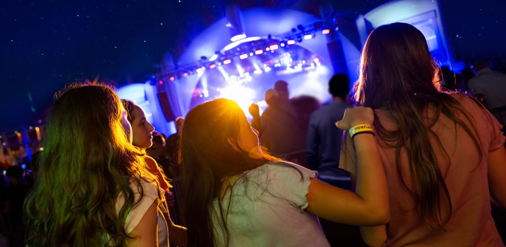 Group of people watching a concert outdoors at night with colorful stage lights and a clear starry sky.