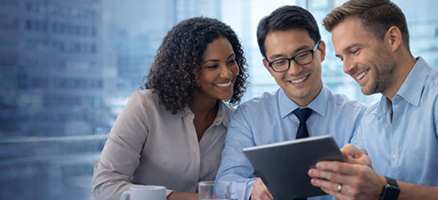 Three smiling professionals looking at a tablet together in a bright office with cityscape view in the background.
