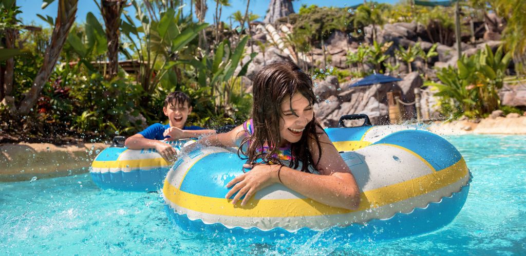 Two children are enjoying a ride on a yellow and blue inflatable tube in a swimming pool with tropical plants and water features in the background.